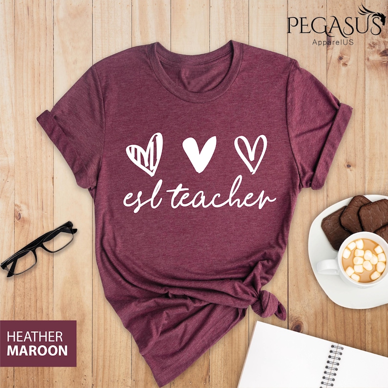 May include: A heather maroon t-shirt with a white graphic of three hearts and the text "esl teacher".