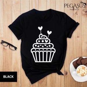 May include: Black t-shirt with a white cupcake design. The cupcake has white hearts and stars on it.