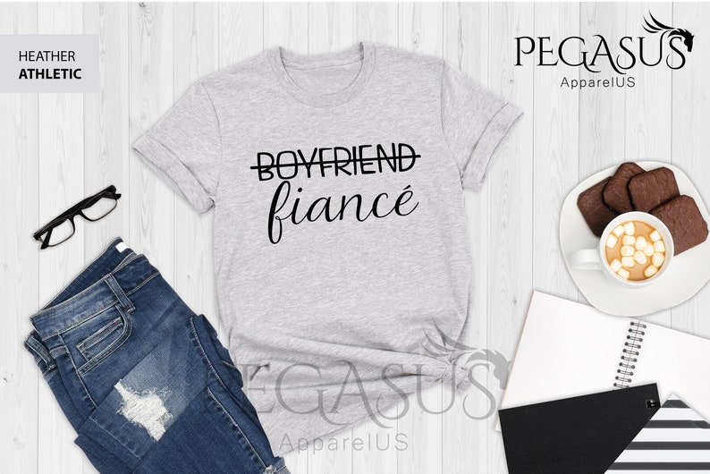 May include: A heather gray t-shirt with the text "BOYFRIEND" crossed out and "fianc&eacute;" written in cursive below it. The shirt is laid flat on a wooden surface with a pair of jeans, a pair of glasses, a cup of coffee, and a notebook.