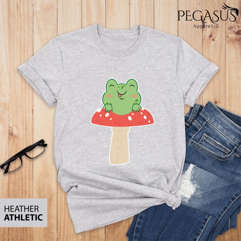 May include: A heather grey t-shirt with a cartoon frog sitting on a red and white mushroom. The frog is green and has a big smile and a tongue sticking out.
