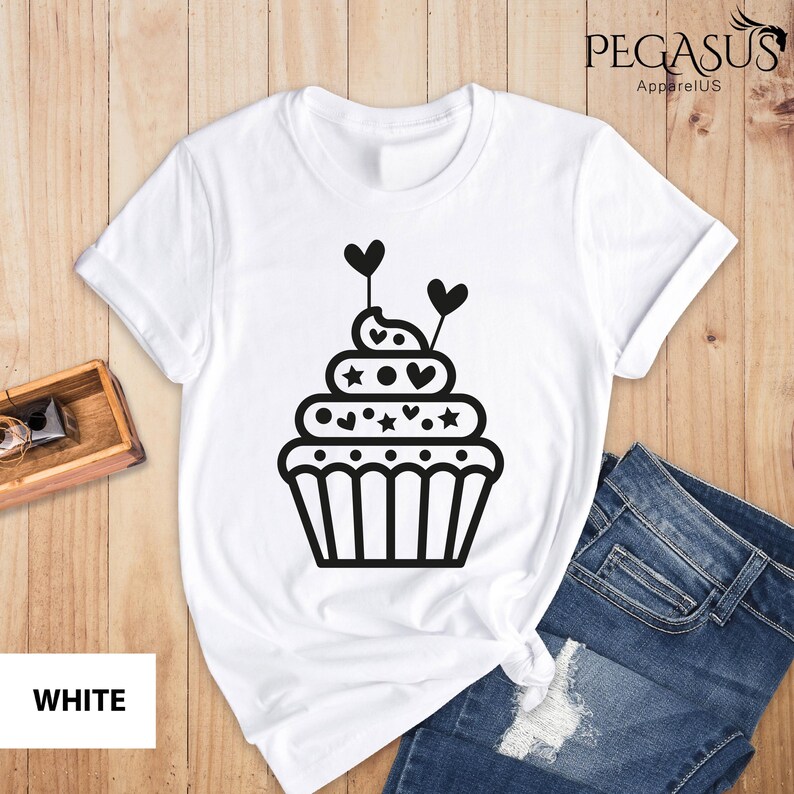 May include: White t-shirt with a black outline of a cupcake with hearts and stars on top. The text "PEGASUS ApparelUS" is in the top right corner of the image.