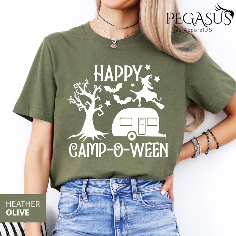 May include: Olive green t-shirt with a white graphic. The graphic includes a tree, bats, a witch on a broom, a camper, and the words "HAPPY CAMP-O-WEEN". The shirt is made of a soft material.