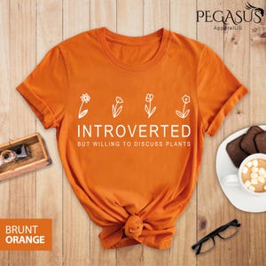 May include: Orange t-shirt with white text that reads "Introverted but willing to discuss plants". The text is surrounded by five small white flower illustrations.