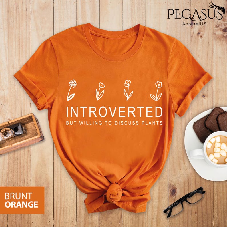 May include: Orange t-shirt with white text that reads "Introverted but willing to discuss plants". The text is surrounded by five small white flower illustrations.