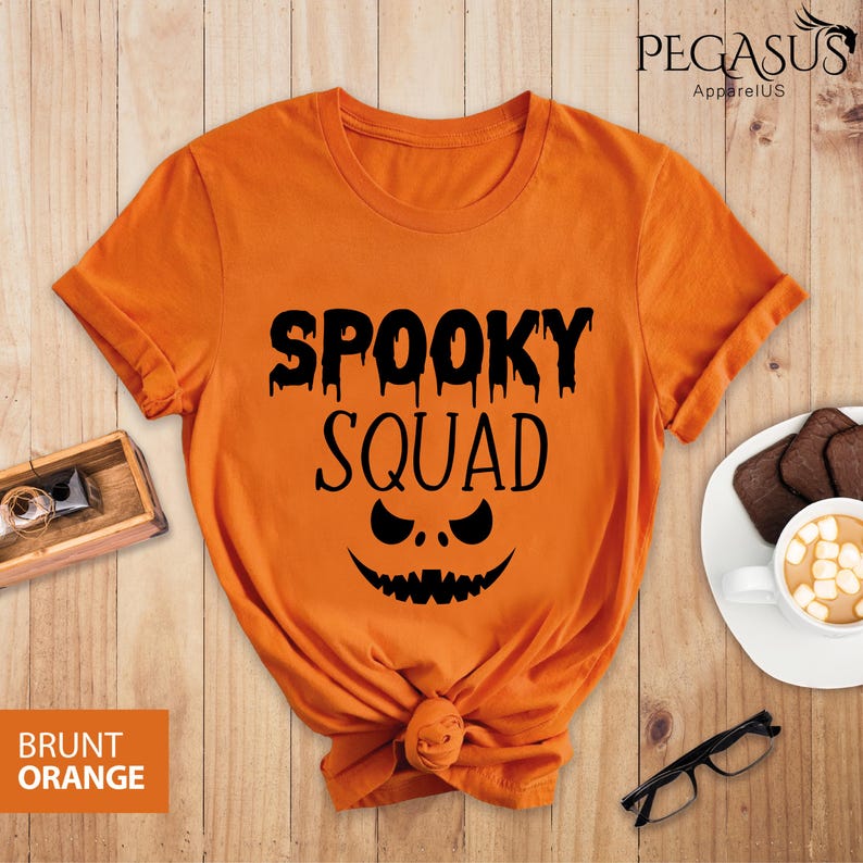 May include: Brunt orange t-shirt with the text "SPOOKY SQUAD" in black, featuring a jack-o'-lantern face design. The shirt is knotted at the bottom. The Pegasus ApparelUS logo is visible in the top right corner.