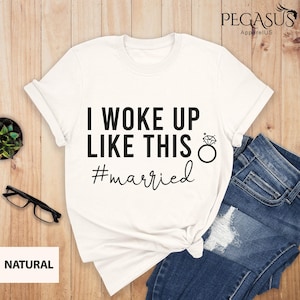 May include: White t-shirt with the text "I WOKE UP LIKE THIS" and "#married" in black. A diamond ring graphic is next to the text. The shirt is paired with blue jeans.