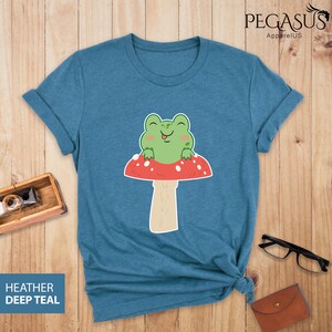 May include: A teal blue t-shirt with a cartoon frog sitting on a red and white mushroom. The frog is smiling and has its tongue sticking out.