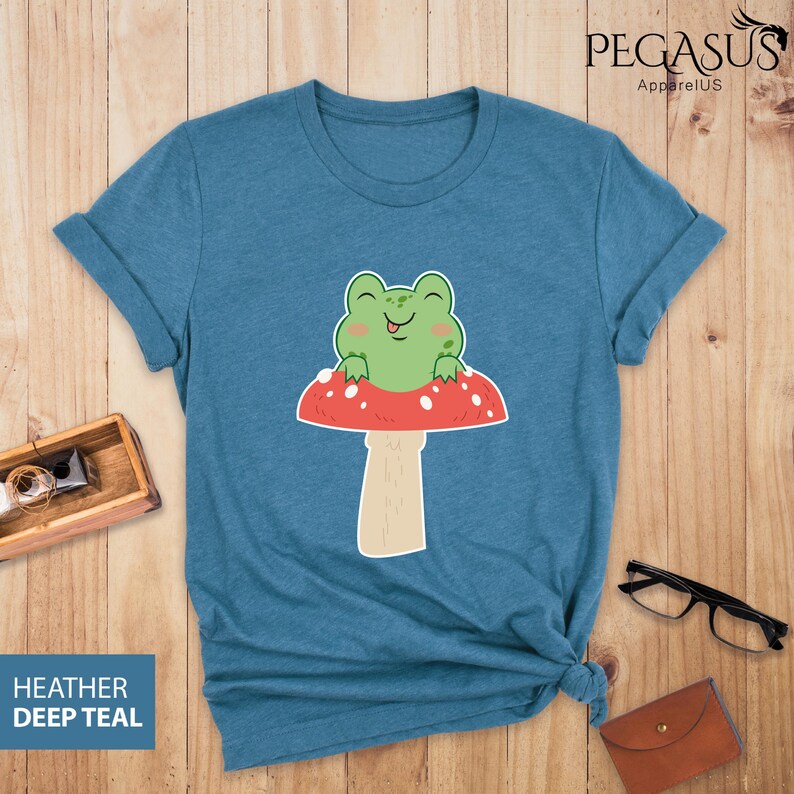 May include: A teal blue t-shirt with a cartoon frog sitting on a red and white mushroom. The frog is smiling and has its tongue sticking out.
