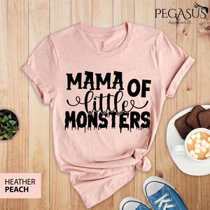 May include: Heather peach t-shirt with the text "MAMA OF little MONSTERS" in black. The shirt is styled on a wooden surface with a cup of hot chocolate, cookies, and blue sneakers.