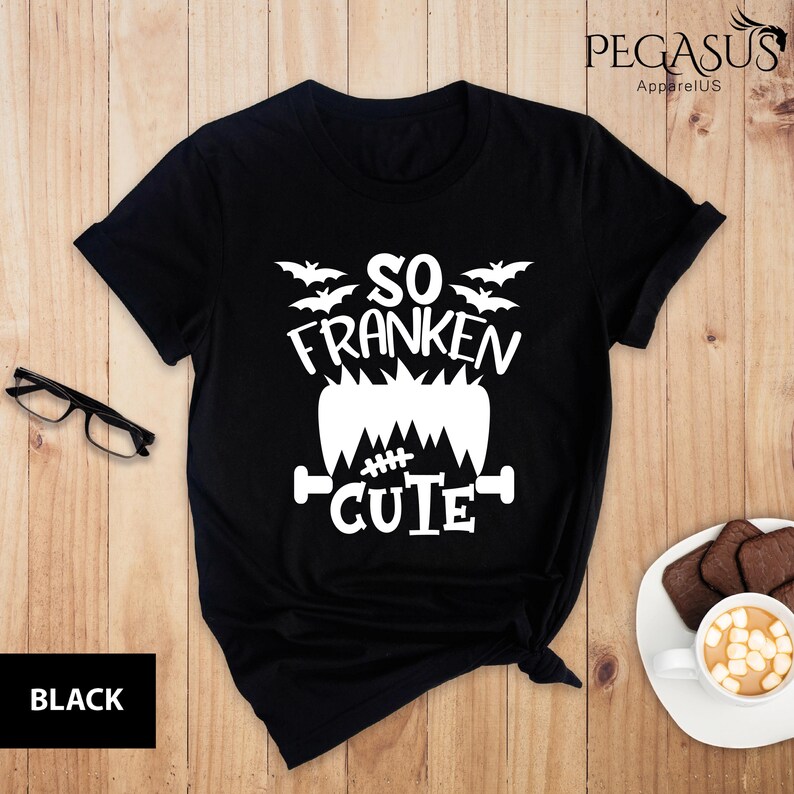 Pu&ograve; includere: T-shirt nero con grafica bianca che dice "SO FRANKEN CUTE" con pipistrelli e un design di mostro di Frankenstein. La maglietta &egrave; di Pegasus ApparelUS.