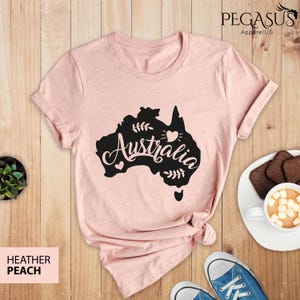 Op de afbeelding: Een roze heather peach t-shirt met een zwarte afbeelding van de omtrek van Australi&euml; met het woord "Australia" in een gestileerd lettertype. De afbeelding bevat ook kleine hartjes en bladeren.