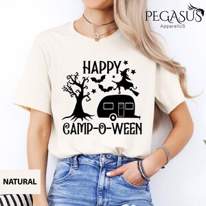 May include: Cream-coloured t-shirt with a black graphic that reads "HAPPY CAMP-O-WEEN." The design includes a tree, bats, stars, a witch on a broom, and a camper. The shirt is made of a soft material.