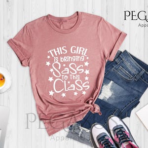K&ouml;nnte beinhalten: Ein T-Shirt in Heide-Mauve mit dem Text "This girl is bringing sass to this class" in wei&szlig;en Buchstaben, umgeben von wei&szlig;en Sternen.