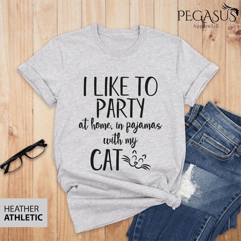 Op de afbeelding: Een grijs t-shirt met de tekst "I LIKE TO PARTY at home, in pajamas with my CAT" in zwarte letters. De kat heeft een zwarte omtrek en een wit gezicht met zwarte ogen en snorharen.
