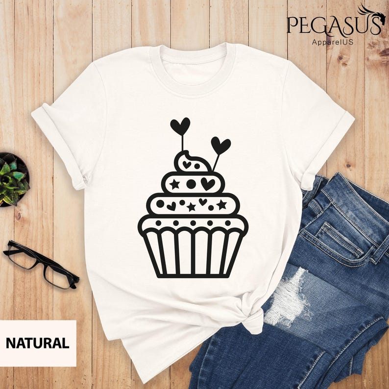 May include: White t-shirt with a black cupcake graphic. The cupcake has hearts and stars on it.