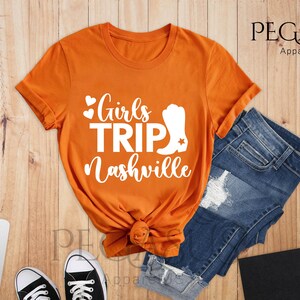 May include: Orange t-shirt with the text "Girls Trip Nashville" in white script and a white cowboy boot graphic. The shirt is paired with blue denim shorts and black sneakers on a wooden surface.