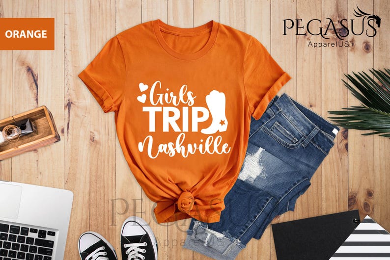May include: Orange t-shirt with the text "Girls Trip Nashville" in white script and a white cowboy boot graphic. The shirt is paired with blue denim shorts and black sneakers on a wooden surface.