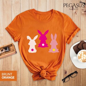 May include: Burnt orange t-shirt featuring three bunny silhouettes in white, pink, and patterned pink. The shirt has a knotted hem and the text "BRUNT ORANGE" is visible. The brand name "PEGASUS ApparelUS" is printed at the top.