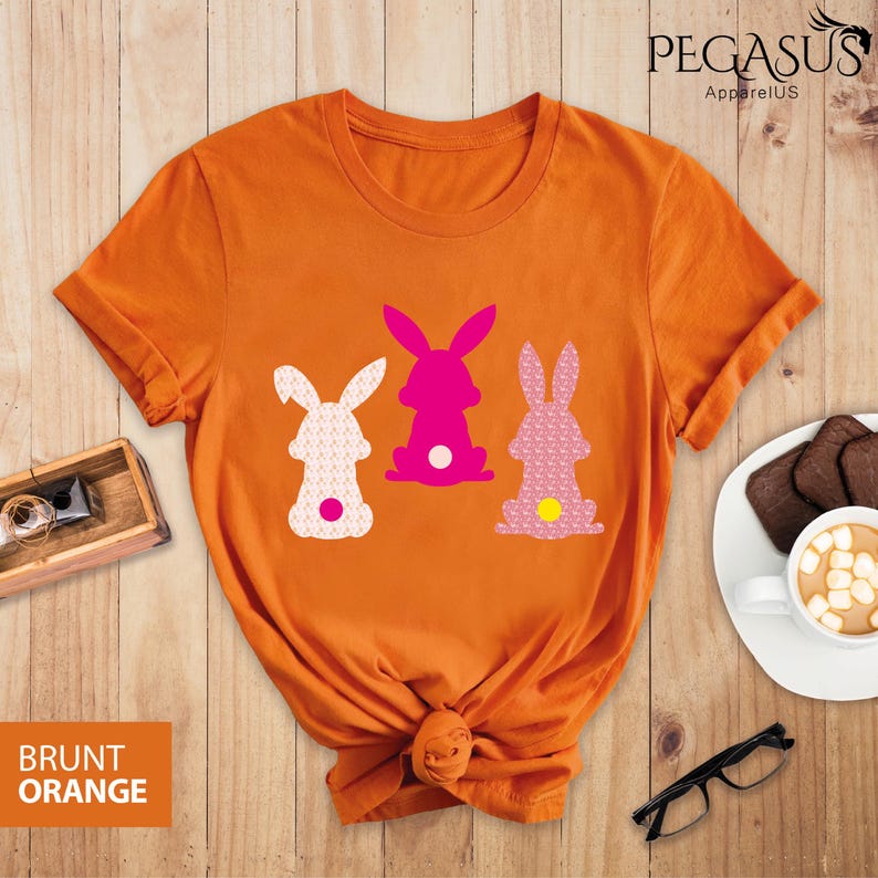 May include: Burnt orange t-shirt featuring three bunny silhouettes in white, pink, and patterned pink. The shirt has a knotted hem and the text "BRUNT ORANGE" is visible. The brand name "PEGASUS ApparelUS" is printed at the top.