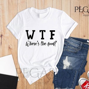May include: White t-shirt with black text that reads "WTF Where's the food?" The shirt is laid out on a wooden surface with a pair of blue jeans and red trainers.