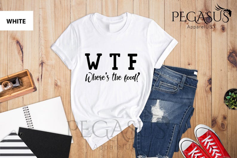 May include: White t-shirt with black text that reads "WTF Where's the food?" The shirt is laid out on a wooden surface with a pair of blue jeans and red trainers.
