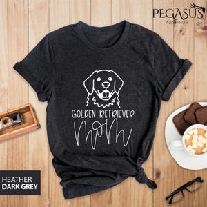 Op de afbeelding: Heather donkergrijs T-shirt met een witte omtrek van een golden retriever en de tekst "Golden Retriever Mom" in wit.