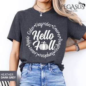 May include: Heather dark grey t-shirt with a white circular design that says "Hello Fall" with a pumpkin graphic. The design also includes the words "Apple, Cider, Hayrides, S'mores, Bonfires, Pumpkins, Leaves".