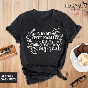 K&ouml;nnte beinhalten: Dunkelgraues T-Shirt mit wei&szlig;em Text, der "Into my craft room I go to lose my mind and find my soul" lautet. Der Text ist von verschiedenen handwerklichen Symbolen umgeben.