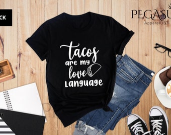 Könnte beinhalten: Schwarzes T-Shirt mit weißem Text, der "Tacos are my love language" lautet, und einer weißen Umrandung eines Tacos. Das Shirt liegt auf einer Holzoberfläche mit einer Jeans, einem Notizbuch und einer Tasse Kaffee.