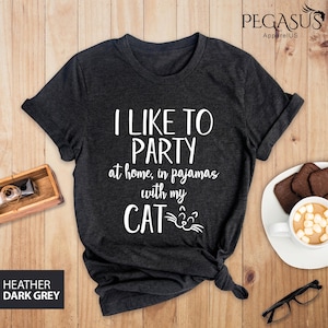 Op de afbeelding: Donkergrijs t-shirt met de tekst "Ik vind het leuk om te feesten thuis, in pyjama met mijn kat" en een cartoon kattengezicht.
