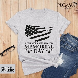 Peut inclure: T-shirt gris chin&eacute; avec un motif de drapeau am&eacute;ricain noir et blanc. Le texte "Remember and Honor Memorial Day" est imprim&eacute; sous le drapeau.