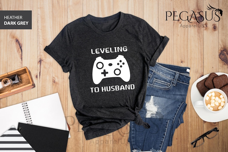 May include: Dark heather grey t-shirt with a white graphic of a video game controller and the text "Leveling to Husband".