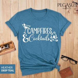 May include: A deep teal t-shirt with the words "Campfires & Cocktails" in white script and a cocktail glass and campfire graphic. The shirt is made of a soft material and has rolled-up sleeves.