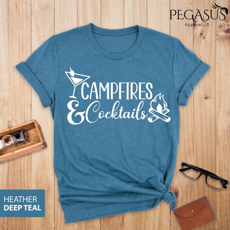May include: A deep teal t-shirt with the words "Campfires & Cocktails" in white script and a cocktail glass and campfire graphic. The shirt is made of a soft material and has rolled-up sleeves.