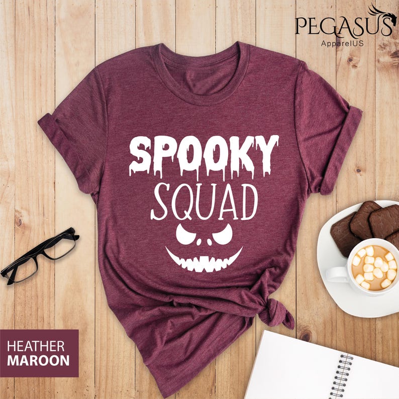 May include: Heather maroon t-shirt with the text "SPOOKY SQUAD" in white, featuring a jack-o'-lantern face design. The shirt is displayed on a wooden surface with a pair of glasses, a notepad, and a cup of coffee.