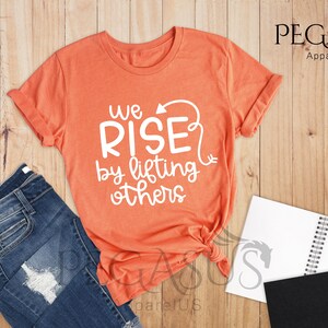 Pu&ograve; includere: Una maglietta color arancio heather con il testo "We Rise by lifting others" in lettere bianche. La maglietta &egrave; annodata in basso.