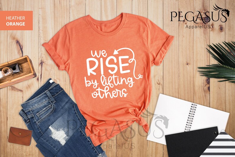 Pu&ograve; includere: Una maglietta color arancio heather con il testo "We Rise by lifting others" in lettere bianche. La maglietta &egrave; annodata in basso.