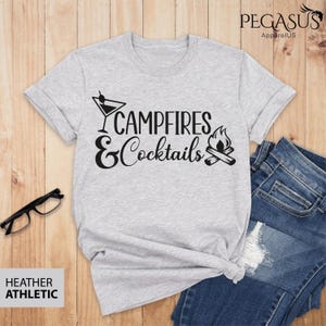May include: Heather athletic gray t-shirt with the text "Campfires & Cocktails" in black. The design includes a martini glass and a campfire graphic. The shirt is paired with blue jeans and black glasses.