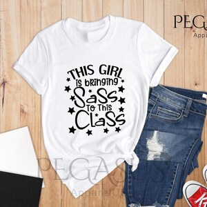 K&ouml;nnte beinhalten: Wei&szlig;es T-Shirt mit schwarzem Text, der "This girl is bringing sass to this class" lautet, umgeben von schwarzen Sternen.