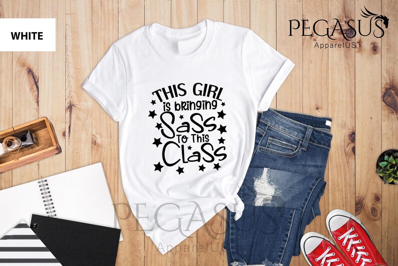 K&ouml;nnte beinhalten: Wei&szlig;es T-Shirt mit schwarzem Text, der "This girl is bringing sass to this class" lautet, umgeben von schwarzen Sternen.