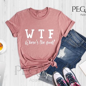 May include: A heather mauve t-shirt with the text "WTF Where's the food?" printed in white on the front. The shirt is laid flat on a wooden surface with a pair of jeans, a laptop, a cup of tea, sunglasses, and a notebook with flowers.