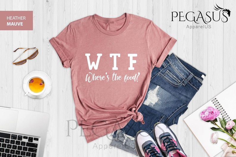 May include: A heather mauve t-shirt with the text "WTF Where's the food?" printed in white on the front. The shirt is laid flat on a wooden surface with a pair of jeans, a laptop, a cup of tea, sunglasses, and a notebook with flowers.