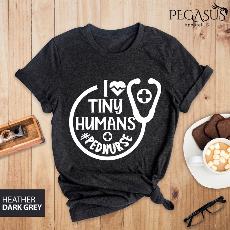 Op de afbeelding: Donkergrijs t-shirt met een wit grafisch ontwerp. De grafiek toont een stethoscoop met een hart en de tekst "Ik hou van kleine mensen #pednurse".