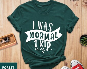 May include: Forest green t-shirt with the text "I WAS NORMAL 1 KID ago" in white. The shirt is folded and has rolled-up sleeves. Red canvas sneakers are visible at the bottom of the image.