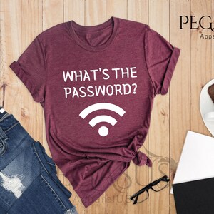 May include: A heather maroon t-shirt with the text "What's the password?" and a white wifi symbol. The shirt is laid out on a wooden surface with a pair of jeans, a notebook, and a cup of hot chocolate.