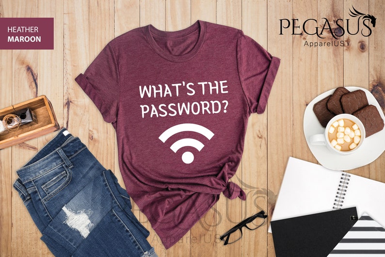 May include: A heather maroon t-shirt with the text "What's the password?" and a white wifi symbol. The shirt is laid out on a wooden surface with a pair of jeans, a notebook, and a cup of hot chocolate.