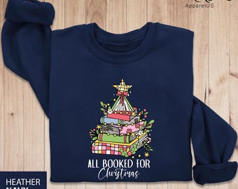 May include: A navy blue sweatshirt with a Christmas tree made of stacked books. The tree is decorated with lights, a star, and greenery. The text "All Booked for Christmas" is printed below the tree.