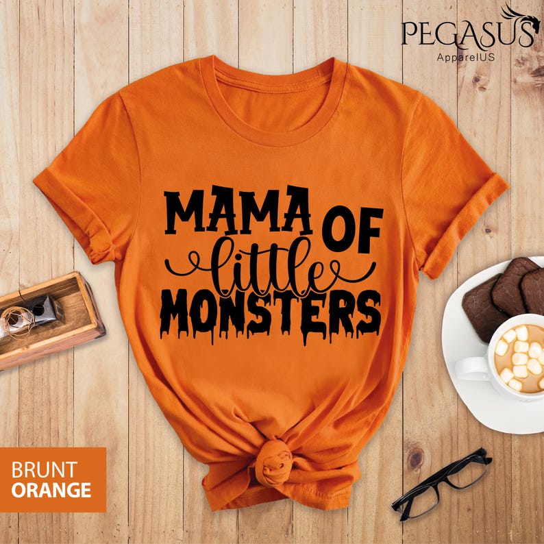 May include: Burnt orange t-shirt with the text "MAMA OF little MONSTERS" in black. The shirt is knotted at the bottom and has short sleeves. The brand name "PEGASUS ApparelUS" is printed at the top right.