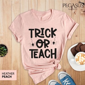 May include: Heather peach-colored t-shirt with the text "TRICK OR TEACH" in black, featuring spider illustrations. The shirt is knotted at the bottom. The image includes a pair of blue sneakers, a plate of cookies, and a cup of hot chocolate.