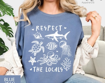 May include: Blue jean colored t-shirt with white graphic of sea creatures and the text "RESPECT THE LOCALS". The design includes a seashell, shark, turtle, fish, jellyfish, starfish, octopus, and coral.